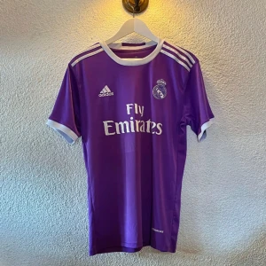 Real Madrid 16/17 Match Kit - Cristiano Ronaldo #7 - Säljer en lila Real Madrid fotbollströja från Adidas med Ronaldo 7 på ryggen. Tröjan har vita detaljer, korta ärmar och klubbmärke samt Fly Emirates-tryck på bröstet. Tillverkad i lätt och ventilerande material, perfekt för fotbollsälskare. Passar S också