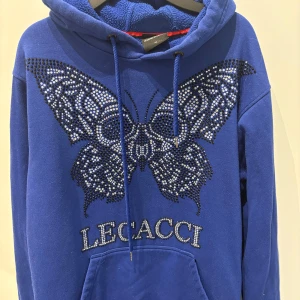 Blå hoodie från Lecacci - Blå hoodie från Lecacci med stor glittrig fjäril i strass på bröstet och texten 'LECACCI' under. Tröjan har huva med snörning, magficka och är fodrad med mjuk fleece.                                                                                (Vissa paljetter har lossnat i tvätten, men syns inte så tydligt)