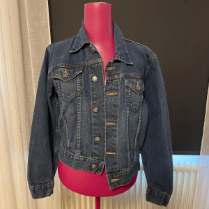 Klassisk jeansjacka från Lipp - Snygg mörkblå jeansjacka från Lipp med klassiska metallknappar och två bröstfickor med lock. Jackan har markerade sömmar i kontrastfärg och en tidlös passform. Passar en s-m. 