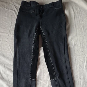 Svarta ridbyxor från Mt. Horse - Svarta ridbyxor från Milton med förstärkta partier på insidan av benen och vita kontrastsömmar. Byxorna har en dragkedjeficka fram och hällor för bälte. Materialet är stretchigt och bekvämt, perfekt för ridning.