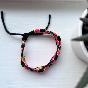 Färgglatt knytarmband i neonfärger - Ett handgjort armband i flätad stil med svarta, rosa och gula neonfärgade trådar. Armbandet har en vågig form och knytstängning som gör det enkelt att justera storleken. Perfekt accessoar för dig som gillar färg och unika detaljer. Aldrig använt! Köpt på Medeltidsveckan på Gotland 🩷 Önskar du veta exakta mått? Tveka inte att höra av dig!