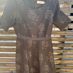 Mörkgrå blommig klänning från Cream - Supersnygg svart klänning från Cream med subtilt blommönster och korta ärmar. Klänningen har rund halsringning och markerad midja för en feminin siluett. Perfekt för dig som gillar stilrena detaljer och blommiga prints.