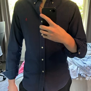 Blå skjorta från Polo Ralph Lauren - Hej, Säljer en mörkblå skjorta från Polo Ralph Lauren i slim fit. Skjortan har klassisk button-down krage, vita knappar och röd broderad logga på bröstet. Tillverkad i mjuk bomull och har långa ärmar. Hör av dig om du har några frågor eller fundering!😁