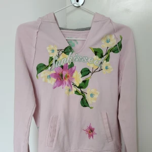 Ljusrosa hoodie med blommor  - Ljusrosa hoodie från EDC by Esprit med broderad text 'Sunkissed' och stora blommiga tryck i gult, grönt och rosa. Tröjan har huva, magficka och lång ärm. Storlek M, men passar också som S.