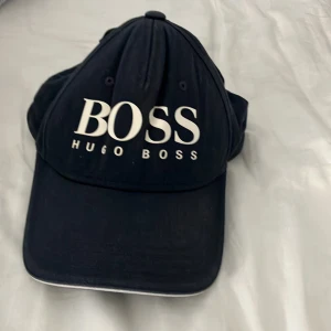 Svart keps från Hugo Boss - Snygg svart keps från Hugo Boss med stor vit logga framtill. Justerbar rem bak med grön detalj och metallspänne med BOSS-gravyr. Insidan har en grön kant som ger extra edge. Perfekt accessoar för en clean och sportig look.