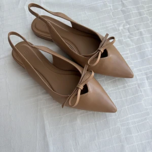 Beige ballerinaskor med rosett från H&M - Stilrena beige ballerinaskor från H&M med spetsig tå och söt rosettdetalj framtill. Skorna har öppen häl med elastiskt band och är tillverkade i syntetmaterial. Perfekta för dig som gillar en chic och enkel look.