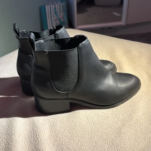 Svarta chelsea boots i skinn - Stilrena svarta chelsea boots med elastiska paneler på sidorna och dragflikar bak. Skorna har låg klack och rund tå, tillverkade i slätt skinn. Perfekta för dig som vill ha en klassisk och clean look till höst och vinter.