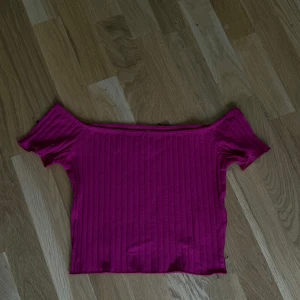 Ribbad offshoulder topp i cerise - Snygg ribbad topp i starkt cerise färg med offshoulder-modell och korta ärmar. Perfekt för dig som vill sticka ut med en färgglad och trendig look. Stretchigt material som sitter skönt och framhäver axlarna.