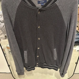 Grå bomberjacka från Gant - Snygg grå bomberjacka från Gant med mörkare grå paneler framtill och baktill. Jackan har ribbade muddar, knappar framtill och två fickor. Materialet är mjukt och känns som bomull, perfekt för en chill och stilren look.