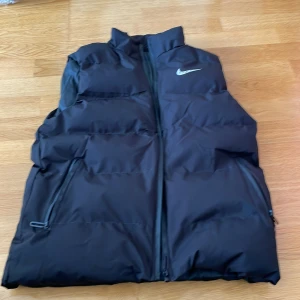 Svart dunväst från Nike - Snygg svart dunväst från Nike med hög krage och dragkedja framtill. Västen har två praktiska fickor med dragkedja och en diskret Nike-logga på bröstet. Perfekt för lager-på-lager och en sportig look.
