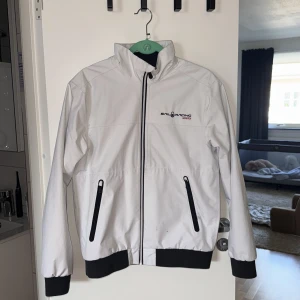 Vit Sail Racing Gore-Tex vindjacka - 🔔Priset kan diskuteras                          Snygg vit vindjacka från Sail Racing med svarta detaljer och dragkedjor. Jackan är tillverkad i slitstark Gore-Tex som håller dig torr. Hög krage, två fickor med dragkedja och svart logga på bröstet. Perfekt för dig som vill ha en clean och sportig look.