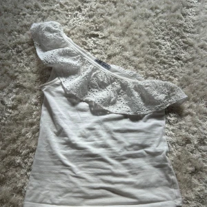 Vit oneshoulder topp med volang - Superfin vit oneshoulder topp från Lindex med bred volang i broderad spets över axeln. Toppen är ärmlös på ena sidan och har en snygg asymmetrisk design. Som ny och inga defekter!
