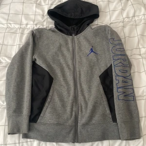 Grå Jordan hoodiejacka med blå logga - Snygg grå hoodiejacka från Jordan med svart huva och svarta detaljer vid sidorna. Stor blå Jumpman-logga på bröstet och JORDAN-tryck längs ena ärmen. Jackan har dragkedja hela vägen och är perfekt för en sportig look.