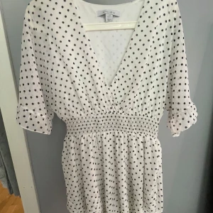 Vit prickig kortklänning från New yorker  - Supersöt vit kortklänning från New yorker med svarta prickar, v-ringad hals och smockad midja. Klänningen har korta ärmar med volangkant och ett luftigt, flowy tyg som känns perfekt för varma dagar.