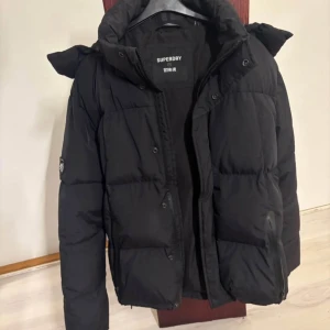 Svart pufferjacka från Superdry stl S - Säljer en svart pufferjacka från Superdry med huva och dragkedja. Jackan har en klassisk boxig passform, stora fickor och diskret logga på armen. Perfekt för kalla dagar och riktigt snygg till streetwear-stilen.