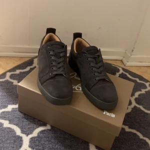 Mörk gråa Louboutins - Snygga mörk gråa sneakers från Christian Louboutin i mocka med klassisk röd sula. Skorna har rund tå, mörk gråa snören och diskret branding på insidan. Insidan är i ljust skinn för extra komfort. Perfekta för dig som vill ha en stilren och lyxig look.