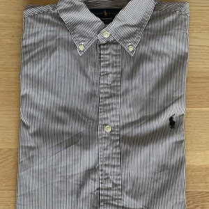 Randig skjorta från Ralph Lauren M - Snygg slim fit skjorta från Ralph Lauren i svartvitt randigt mönster. Klassisk krage, knäppning framtill och broderad logga på bröstet. Tillverkad i 100% bomull för en skön känsla och stilren look.