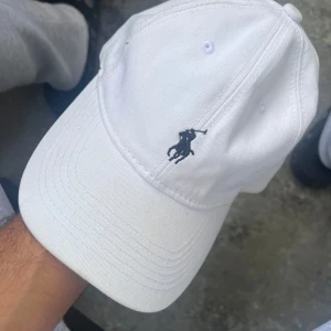 Vit keps från Polo Ralph Lauren - Vit keps från Polo Ralph Lauren med klassisk broderad logga framtill och justerbart spänne bak. Kepsen är i bomull och har snygg, stilren design med Polo-broderi även baktill och på insidan. Perfekt accessoar för en clean och sportig look.