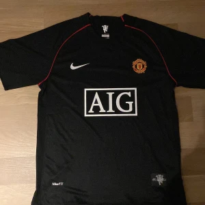 Manchester United Ronaldo 7 Nike tröja - Säljer en svart Manchester United fotbollströja från Nike med Ronaldo 7 på ryggen. Tröjan har röda detaljer, klubbmärke på bröstet och AIG som sponsor framtill. Tillverkad i Nike FITDRY-material som andas. Perfekt för dig som älskar fotboll och United.