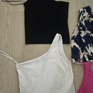 Fyra trendiga one shoulder toppar - Säljer fyra snygga one shoulder toppar: en svart, en vit ribbad, en beige med marinblått blommönster och en knallrosa. Perfekta för dig som vill sticka ut med coola färger och mönster. Alla ärmlösa och i mjuka material, vissa med tunna axelband. Från Zara, H&M 