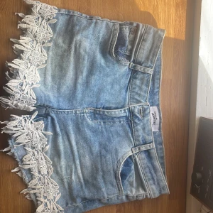 Ljusblå jeansshorts med spets från Jennyfer - Supersnygga ljusblå jeansshorts från Jennyfer med vita spetsdetaljer längs bensluten. Klassisk femficksmodell, bälteshällor och en riktigt somrig vibe. Perfekta för dig som vill sticka ut lite extra med coola detaljer!