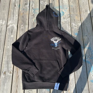 Svart Hoodie från Hoodrich med ängeltryck - Svart hoodie från Hoodrich med stor vit och blå ängelgrafik och texten 'From Nothing To Something' på bröstet, ryggen och huvan. Klassisk känguruficka och ribbade muddar. Skön streetwear vibe, perfekt för dig som gillar snygga detaljer och statement-plagg.