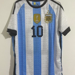 Argentina Messi fotbollströja blåvit - Säljer en officiell Argentina fotbollströja från Adidas med Messi och nummer 10. Tröjan har klassiska blå och vita ränder, svarta detaljer vid ärmslut och krage samt FIFA 2022-märke och AFA-logga på bröstet. Tillverkad i lätt och ventilerande material. Tröjan är helt ny och aldrig använt. 