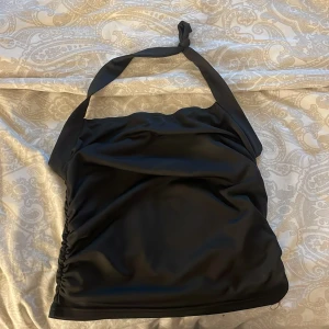 Svart halterneck topp med rynk - Säljer en svart halterneck topp med knyt i nacken och snygg rynkdetalj på sidan. Toppen är ärmlös och har en slät, glansig yta som ger en cool look. Perfekt för dig som gillar stilrena och trendiga plagg.