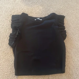 Svart volangtopp från Zara - Säljer en svart topp från Zara med snygga volangdetaljer på axlarna. Modellen är ärmlös och har en rak passform. Perfekt för dig som vill ha en enkel men ändå trendig look. Materialet känns mjukt och följsamt.