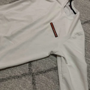 Vit sweatshirt från Prada - Vit sweatshirt från Prada med rund halsringning och diskret logga i rött och svart på bröstet. Tröjan har långa ärmar och ribbade muddar vid hals, ärmslut och nederkant. Enkel och clean design som passar till allt. äkta, finns scan kod. priset kan diskoteras