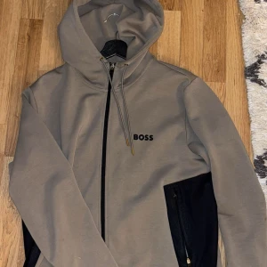 Beige hoodie från BOSS med dragkedja - Snygg beige hoodie från BOSS med svart dragkedja och svarta detaljer på fickorna. Tröjan har huva med snörning och BOSS-logga broderad på bröstet. Perfekt för en chill och stilren look.