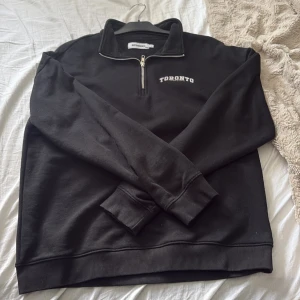 Svart half zip sweatshirt Toronto - Svart sweatshirt från New Garments med broderad text 'Toronto' på bröstet. Tröjan har en klassisk half zip-dragkedja, hög krage och långa ärmar. Mjuk bomullskänsla och relaxed fit, perfekt för chill dagar eller när du vill ha en clean look.