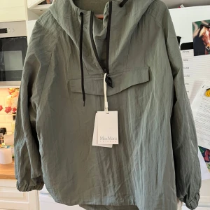 Grön vindjacka från MaxMara Leisure - Snygg grön vindjacka från MaxMara Leisure med huva, dragkedja framtill och justerbara snören. Jackan har en avslappnad passform och elastiska muddar vid ärmsluten. Perfekt för dig som gillar stilren och funktionell design.