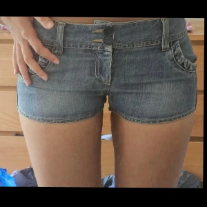 Blå lowwaist jeansshorts - Snygga blå jeansshorts från D. JAWIN 509 med låg midja och dubbla knappar framtill. Shortsen har coola rynkade detaljer på både fram- och bakfickor samt klassiska bälteshällor. Perfekta för sommaren och riktigt sköna i mjukt denimtyg.