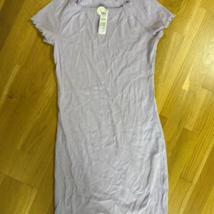 Ljuslila ribbad kortklänning H&M - Supersöt ljuslila ribbad kortklänning från H&M med volangkant vid halsringning och ärmslut. Klänningen har korta ärmar och en figurnära passform som känns både trendig och bekväm. Perfekt för dig som gillar en enkel men ändå söt stil. Helt ny 