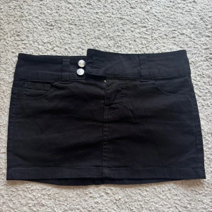 Svart jeanskjol med fickor - Svart kort jeanskjol med två silvriga knappar framtill, klassiska jeansfickor både fram och bak samt hällor för bälte. Snygg och enkel modell som passar till många olika stilar.