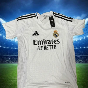 Real Madrid Fotbollströja  - Snygg vit Real Madrid fotbollströja från Adidas med svarta detaljer och klubbmärke på bröstet. Tröjan har korta ärmar, klassiska svarta ränder på axlarna och meshpaneler på sidorna för extra ventilation. Tillverkad i lätt och funktionellt material.
