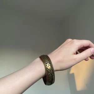 Brett armband i antikguld med mönster - Snyggt brett armband i antikguld med utskuret, blommigt mönster från Indien. Armbandet har en cool vintagekänsla och passar perfekt för dig som gillar unika accessoarer med lite boho-vibe.