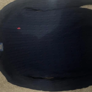 Mörkblå stickad tröja Polo Ralph Lauren - Kabelstickad mörkblå tröja från Polo Ralph Lauren med klassisk röd broderad logga på bröstet. Tröjan har rund halsringning och långa ärmar, perfekt för lager-på-lager under kyliga dagar.