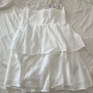 Vit volangklänning - Superfin vit klänning från Gina tricot med smock upptill utan axelband. Klänningen har flera lager volanger och är tillverkad i ett lätt, luftigt bomullstyg. Perfekt för varma dagar när du vill känna dig både söt och bekväm.