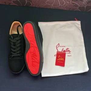 Christian Louboutin svarta sneakers - Exklusiva svarta sneakers från Christian Louboutin med klassisk röd sula. Skorna har rund tå, svarta snören och är tillverkade i mocka med detaljer i skinn. Snygg och stilren design som sticker ut med den ikoniska röda undersidan.