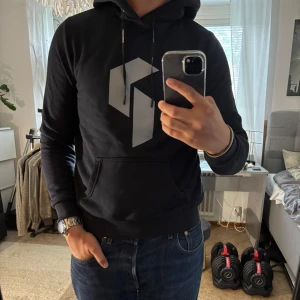 Svart hoodie Peak Performance M - Svart hoodie från Peak Performance med stor grå logga på bröstet. Klassisk känguruficka, snörning i huvan och ribbade muddar. Skön och stilren modell i bomullsmix, perfekt för chill eller träning.