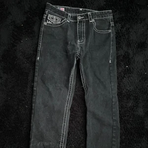 Svarta True Religion jeans med kontrastsömmar - Svarta jeans från True Religion med tydliga vita kontrastsömmar och ikoniska bakfickor med stora broderier. Klassisk femficksmodell i bomull med knappgylf och detaljerade nitar. Perfekta för dig som vill sticka ut med en streetig vibe.