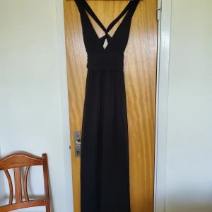 Svart långklänning med korsade band - Elegant svart långklänning med djup v-ringning och breda korsade axelband i ryggen. Klänningen har markerad midja och ett flytande, lätt transparent tyg som ger en snygg siluett. Perfekt för bal eller festkvällar. Etiketten är bortklippt men storlek mellan 36-38. Bra skick! 