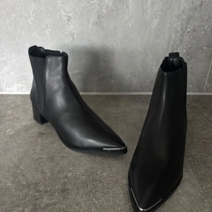 Svarta chelsea boots från Acne Studios - Snygga svarta chelsea boots från Acne Studios i äkta skinn. Skorna har spetsig tå, låg klack och elastiska paneler på sidorna för enkel påtagning. Stilren design med metall-detalj framtill på tån. Perfekta för dig som gillar minimalistisk och edgy stil.