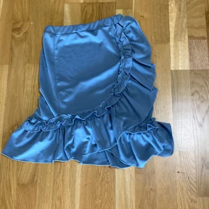 Damm Blå volangkjol med omlottkänsla - Supersöt damm blå kortkjol med volanger och omlottkänsla. Kjolen har en mjuk, flowy look och är perfekt för dig som gillar en feminin och trendig stil. Materialet känns lätt och svalt, och volangdetaljerna ger extra rörelse.