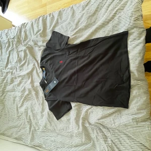 Svart t-shirt från Polo Ralph Lauren - Svart t-shirt från Polo Ralph Lauren med klassisk passform och rund hals. Ikoniska röda Polo-loggan broderad på bröstet. Tillverkad i mjuk bomull, perfekt för en clean och stilren look.