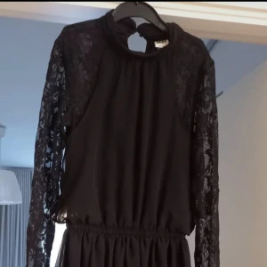 Svart klänning med spetsärmar Vero Moda - Svart långklänning från Vero Moda i stl XS med transparenta spetsärmar och hög hals med knappdetalj.                                               Klänningen har markerad midja med resår och ett lätt, flödande tyg i kjolen.                         Perfekt för dig som gillar en elegant och stilren look.                                                                                              Aldrig använd!