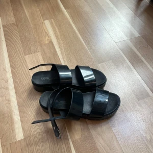 Svarta sandaler från ASOS med breda remmar - Snygga svarta sandaler från ASOS med två breda, blanka remmar och justerbar rem runt hälen. Platt sula och öppen tå ger en clean och modern look. Perfekta för varma dagar när du vill ha både stil och komfort.