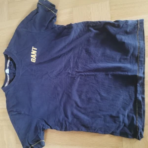 Blå GANT t-shirt med tryck - Mörkblå t-shirt från GANT med gul text på bröstet och stort 'GANT SPORT'-tryck på ryggen. Klassisk passform, rund hals och korta ärmar. Tillverkad i mjuk bomull, perfekt för en sportig och avslappnad stil. 
